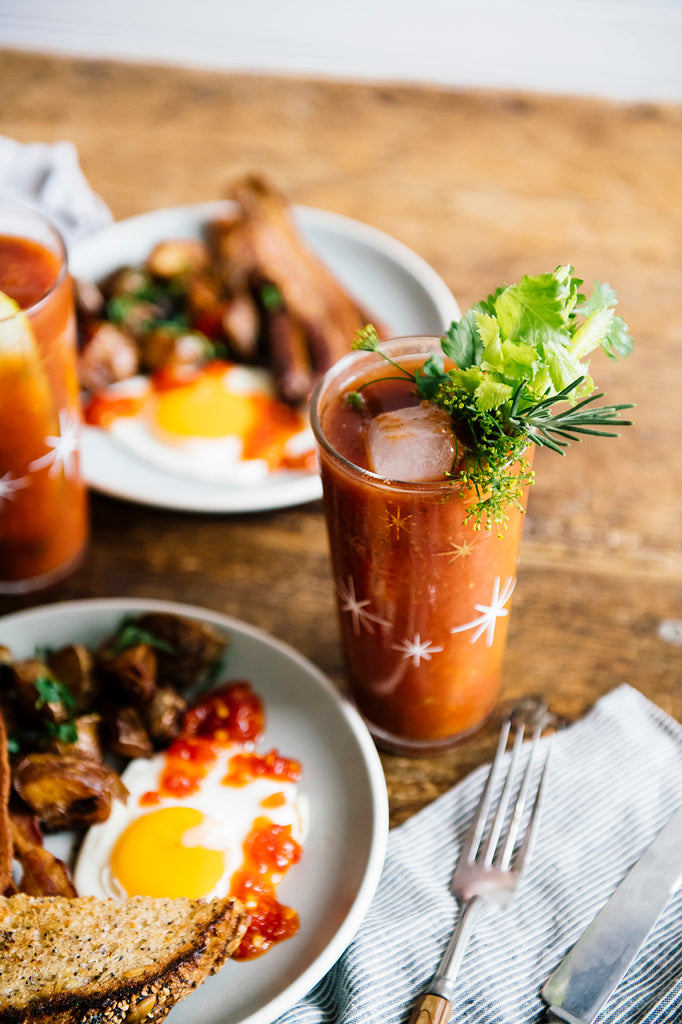 a homemade bloody mary with fresh garnishes next to a breakfast plate of potatoes, eggs, toast and bacon