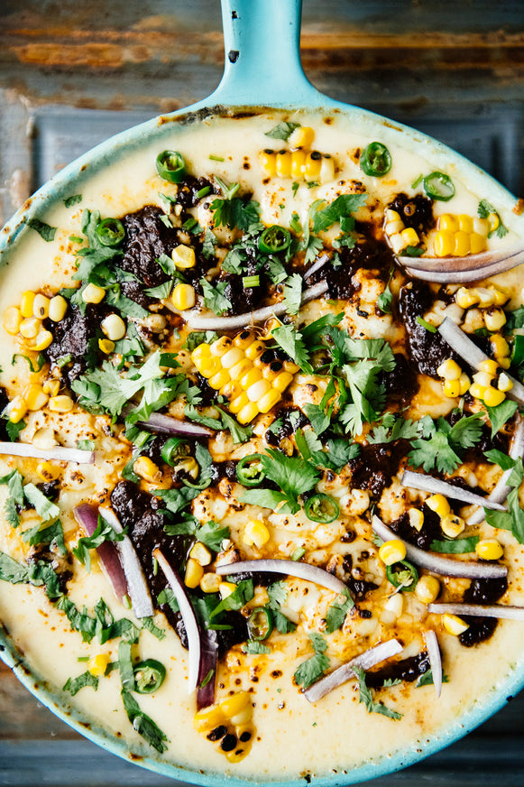 A skillet of melty queso dip topped with sweet corn, slivers of red onion, cilantro, and Dried Chile Salsa