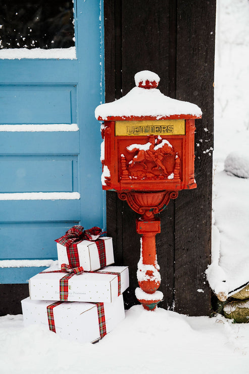 Load image into Gallery viewer, an antique red letter box in the snow with gifts tied with plaid ribbon set next to it in front of a blue door