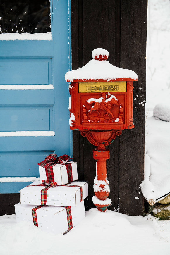 an antique red letter box in the snow with gifts tied with plaid ribbon set next to it in front of a blue door