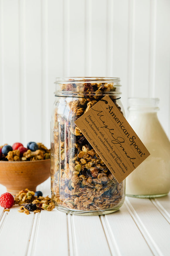 a jar of maple granola with a bowl of granola and berries and a bottle of milk in the background