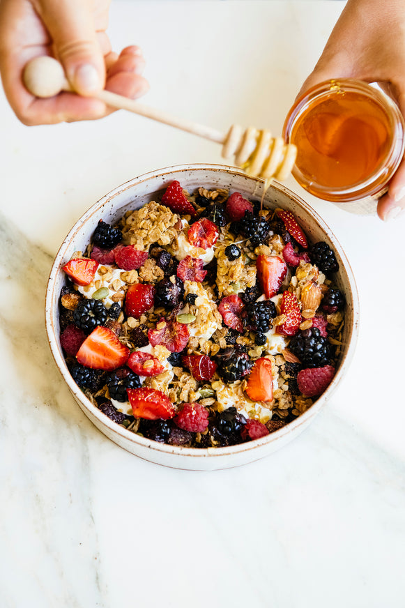 a bowl of granola with fresh berries, with a hand drizzling honey from a honey dipper