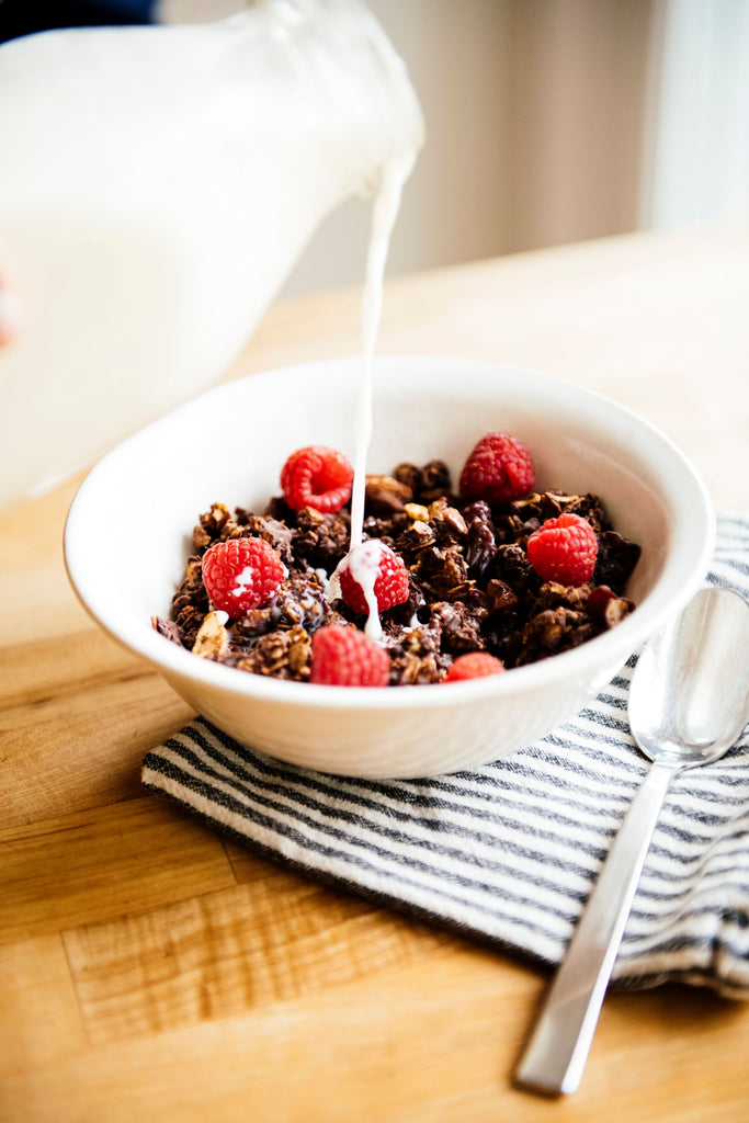 milk being poured over a bowl of chocolate granola topped with fresh raspberries