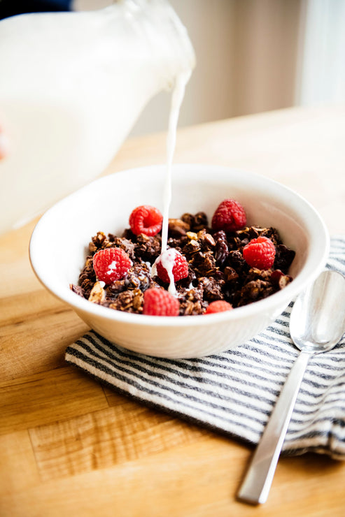 Load image into Gallery viewer, milk being poured over a bowl of chocolate granola topped with fresh raspberries