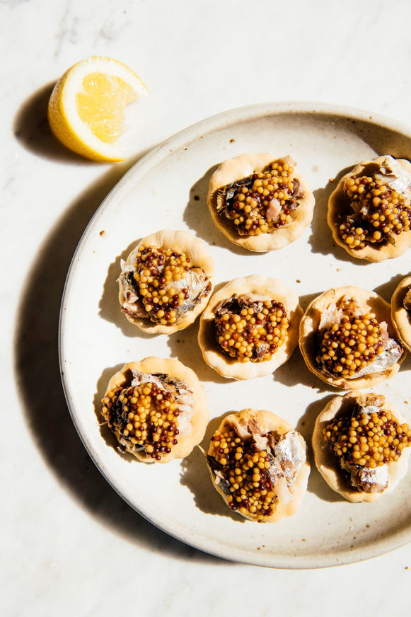a plate of crackers topped with sardines and whole seed mustard with a wedge of lemon