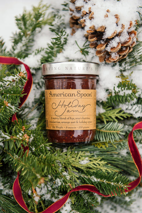 Load image into Gallery viewer, A jar of American Spoon Holiday Jam styled in a snowy pine setting with a red ribbon and a pinecone. The label reads 'American Spoon Holiday Jam' and 'A tasting blend of pine, nut, citrus, and holiday spices'.