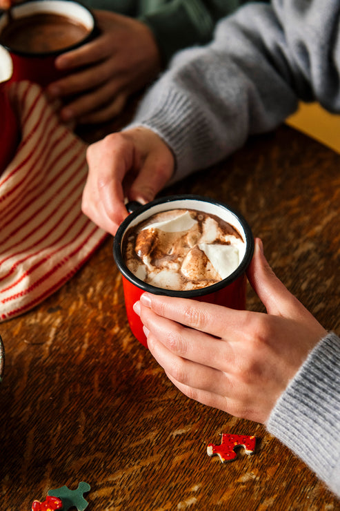 Load image into Gallery viewer, Person holding a mug of hot chocolate with marshmallows on a wooden table.