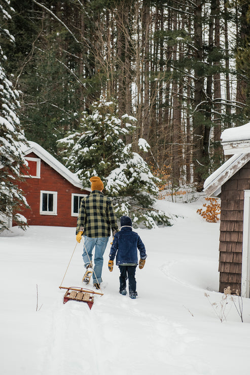 Load image into Gallery viewer, An adult and a child walk through deep snow toward a red cabin, with the man pulling a small wooden sled. Snow-covered evergreen trees and a forest fill the background.