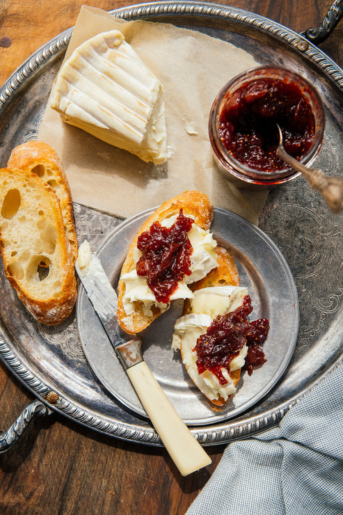 Load image into Gallery viewer, An overhead styled food scene on a large silver tray, showing a slice of white cheese, a jar American Spoon Holiday Jam with a spoon, two pieces of baguette, and two open-faced bread slices topped with cream cheese and the jam, with a knife resting on the tray.