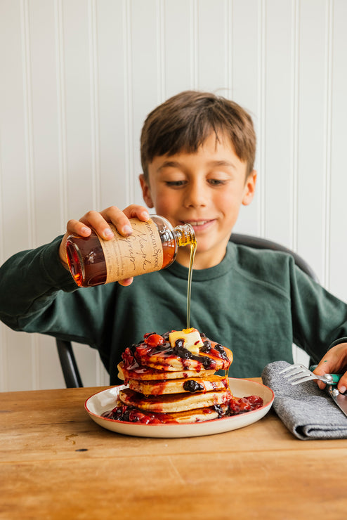 Load image into Gallery viewer, a young boy drizzling maple syrup on top of a stack of pancakes topped with cherry and blueberry fruit perfect