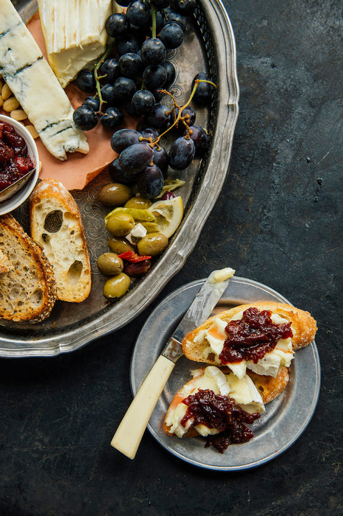 Load image into Gallery viewer, An overhead food scene showing a large silver platter with assorted cheeses, dark grapes, bread slices, olives, and a small bowl of Holiday Jam, plus a smaller plate with bread topped with cheese and red jam and a knife on a dark textured tabletop.