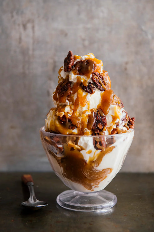 Load image into Gallery viewer, Dessert with ice cream, caramel, and pecans in a glass bowl on a textured surface.