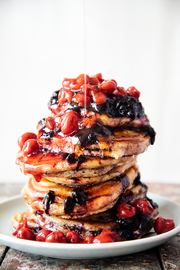 a stack of pancakes topped with maple syrup, fruit perfect sour cherries and fruit perfect blueberries