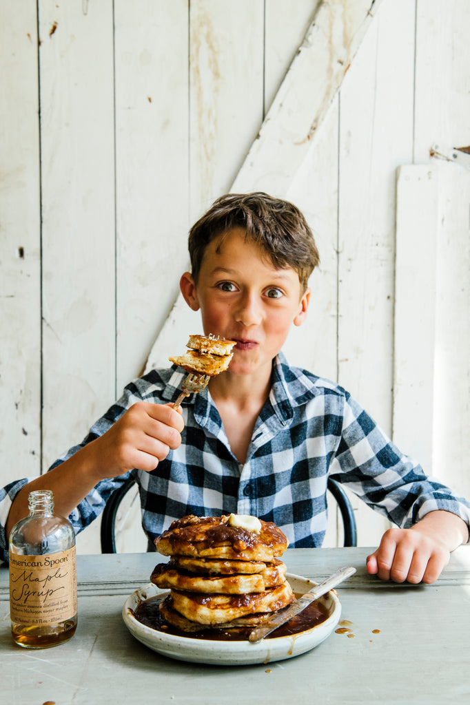 A young boy with a fork full of pancakes and a stack of pancakes covered in Maple Syrup