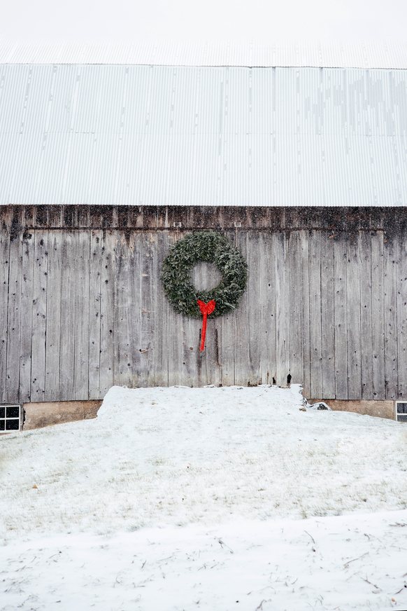 A snow covered barn with a large Christmas wreath and red ribbon