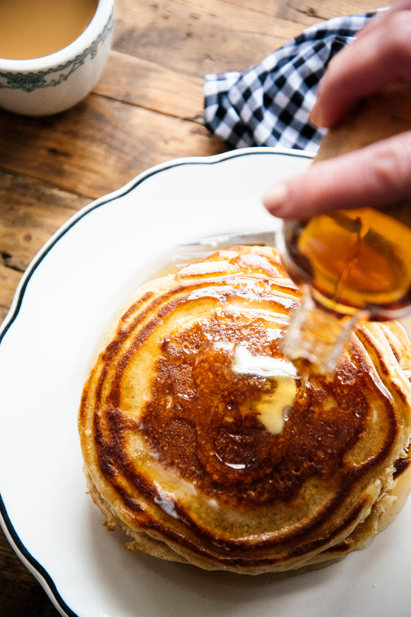 A stack of pancakes with melted butter, and a person pouring maple syrup over them from a small glass bottle.