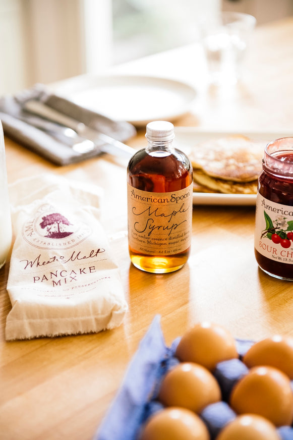 Bottle of maple syrup, pancake mix, Fruit Perfect Sour Cherries and eggs on a table  with blurred pancakes, a plate and silverware in the background