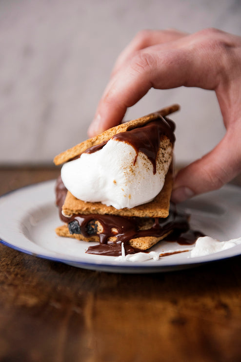 Load image into Gallery viewer, Hand holding a s'mores dessert on a white plate with a blurred background
