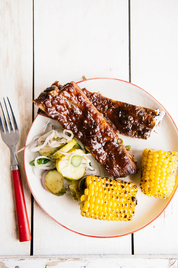 Ribs, corn on the cob and pickles on a red-rimmed white plate.