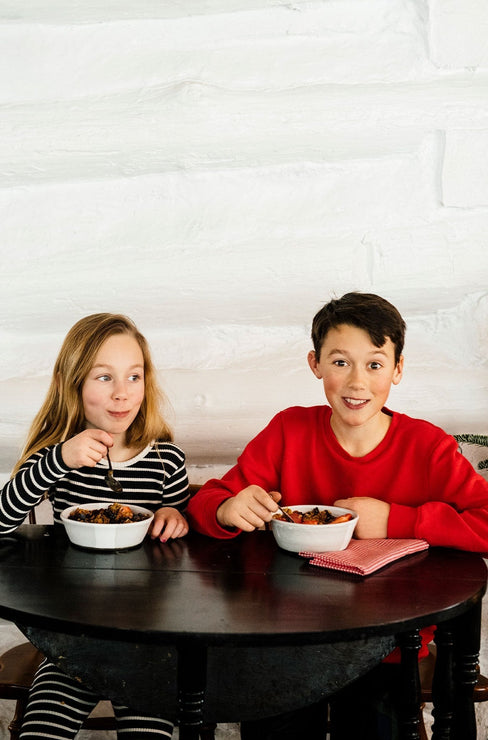 Load image into Gallery viewer, two young children in holiday pajamas eating granola from bowls at a black table
