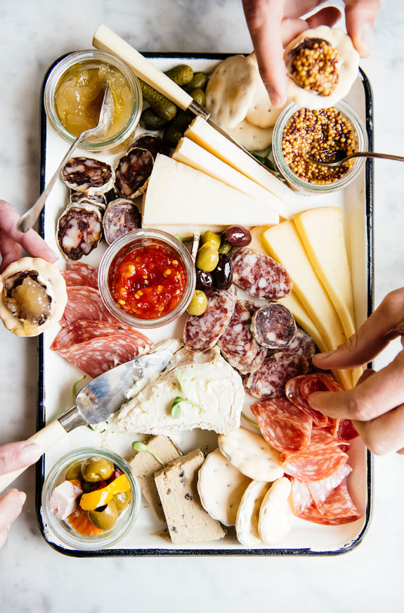 A charcuterie board with hands reaching in to select cheese, sausages.