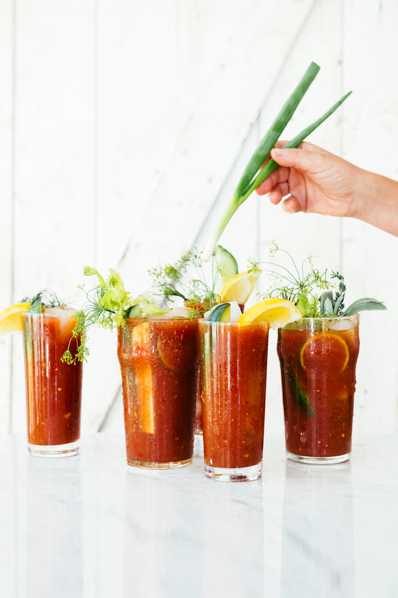 Bloody mary cocktails with herbs and lemon; and a woman's hand about to add a green onion garnish.