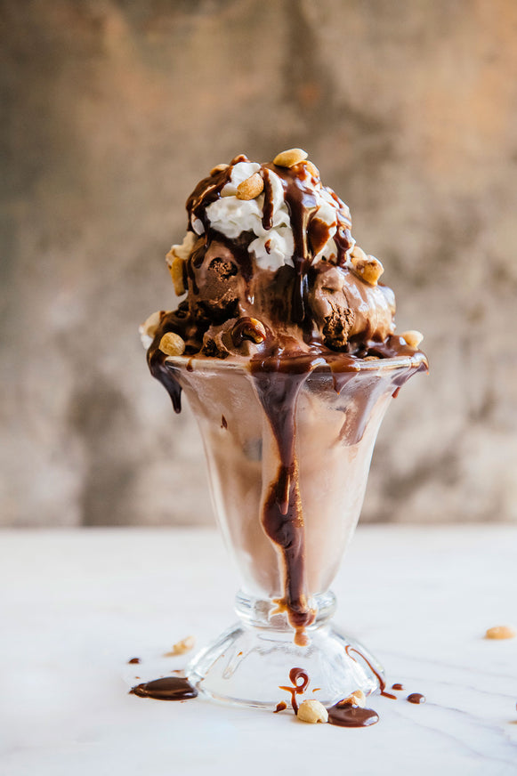 Chocolate ice cream sundae with nuts & whipped cream in a glass cup on a white surface.