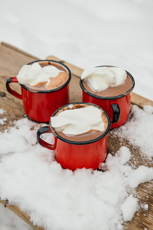 Load image into Gallery viewer, Three red enamel mugs filled with hot cocoa and topped with whipped cream, arranged on a wooden tray surrounded by snow.
