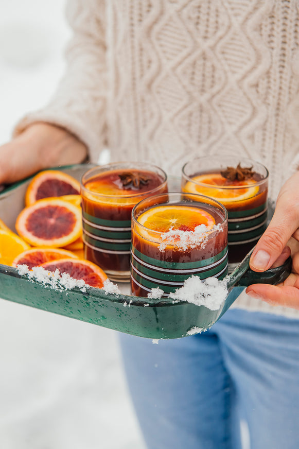 A person in a cream cable-knit sweater and blue jeans holds a dark green tray in a snowy outdoor setting. The tray holds three stacked clear glasses filled with a warm red drink, garnished with orange slices and coarse salt, with additional orange slices on the tray.