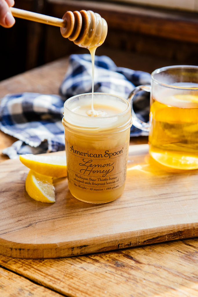 Honey being drizzled out of a jar of lemon honey with tea and lemon slices in the background