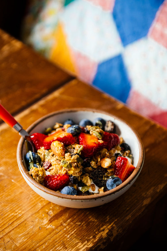 A bowl of granola and berries on a bedside table, next to a bed made with a vintage quilt.