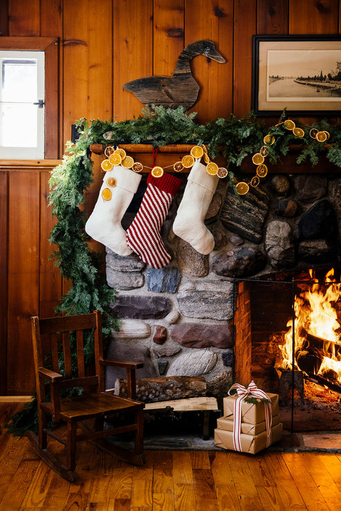 Load image into Gallery viewer, A cozy rustic fireplace scene with three Christmas stockings hanging on a stone mantel decorated with green garland and orange slice garlands. A small wooden rocking chair sits on the hardwood floor beside a wrapped gift box, with a fire burning in the fireplace and wood-paneled walls behind.
