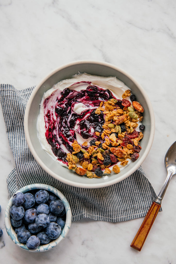 a bowl of yogurt and granola topped with blueberry preserves next to a small bowl of blueberries