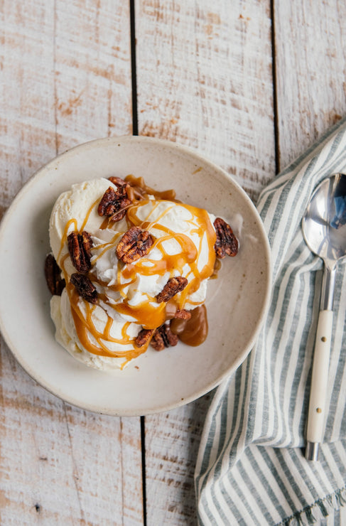 Load image into Gallery viewer, Ice cream with caramel and pecans on a white bowl, placed on a wooden surface with a striped cloth and spoon.
