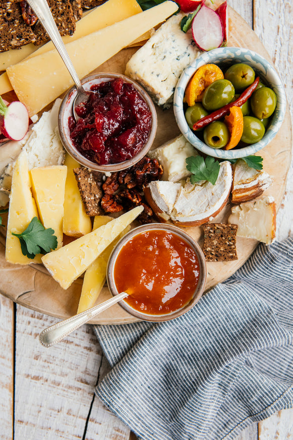 An overhead styled food scene on a wooden board with cheese wedges, crackers, olives, and two small bowls of  jam, set on a rustic white wood table with a gray cloth.