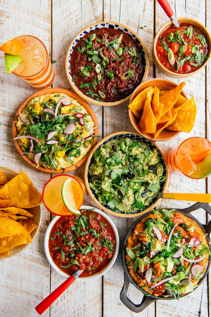 bowls of guacamole, chips, salsa, bean dip, street corn dip and glasses of margaritas