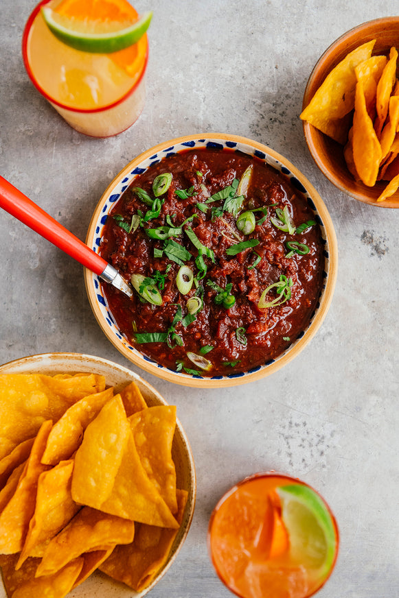 Bowl of Cherry Chipotle Salsa sprinkled with green onions next to bowls of tortilla chips and glasses of margaritas.