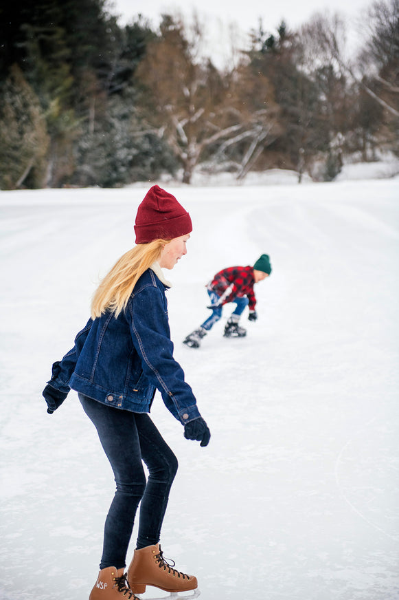 Girl in a ed beanie and blue jacket skating on a snowy ice rink.