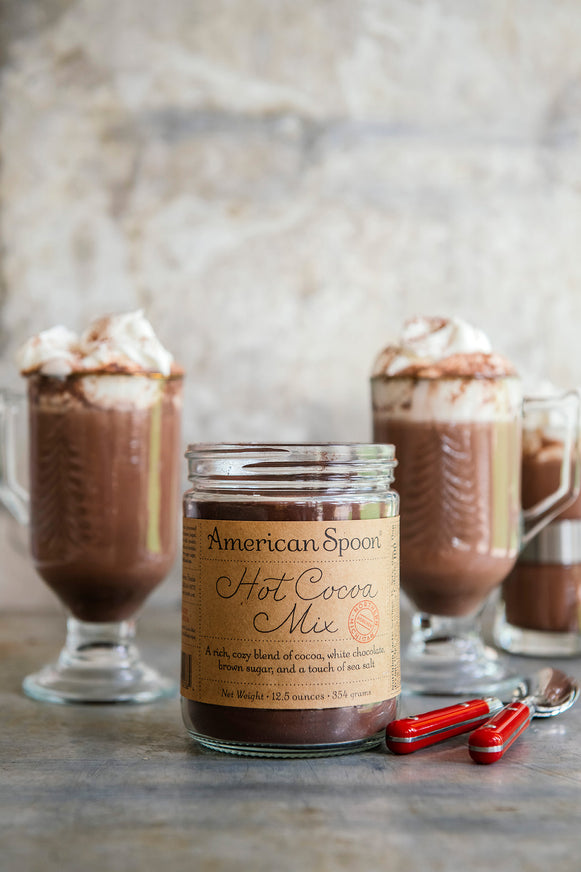 an open jar of hot cocoa mix surrounded by mugs of hot cocoa and fresh whipped cream