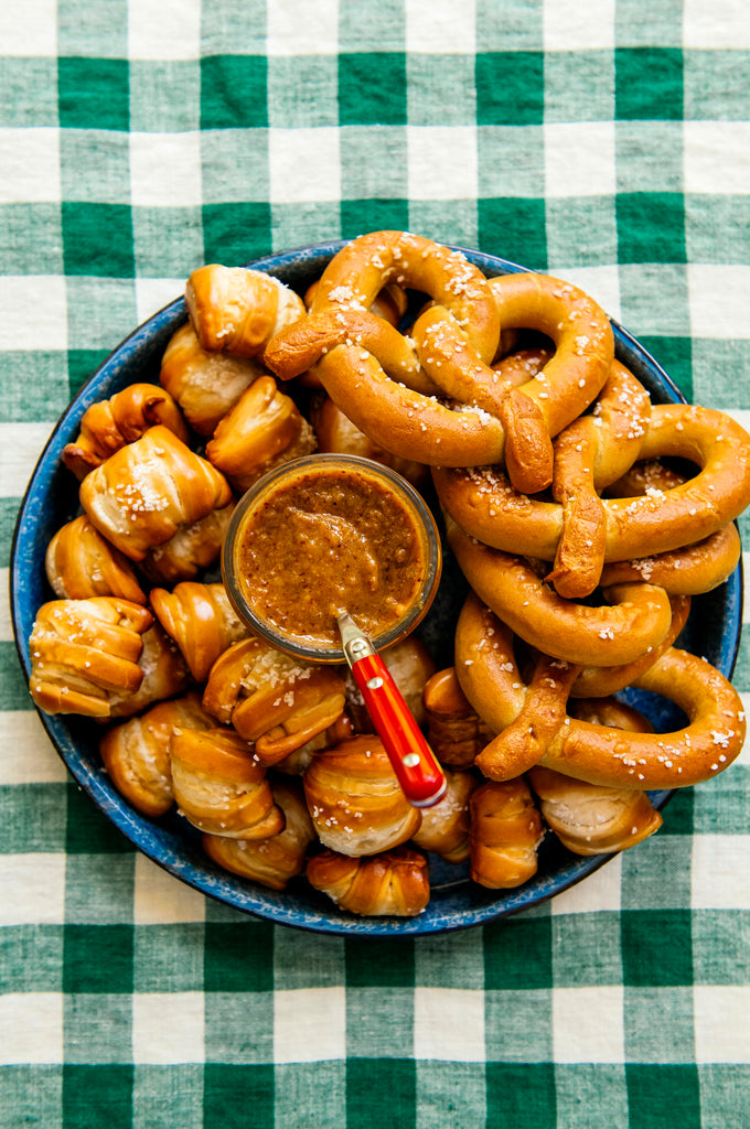 A blue circular tray with soft pretzels and golden ale mustard, set on a green and white checked tablecloth.