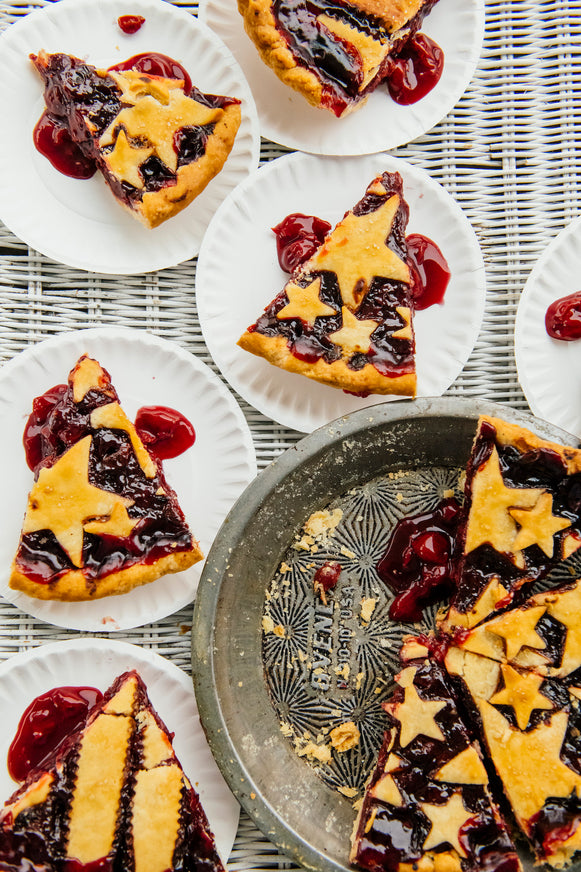 Slices of cherry pie with crust stars on white plates on a white rattan table.