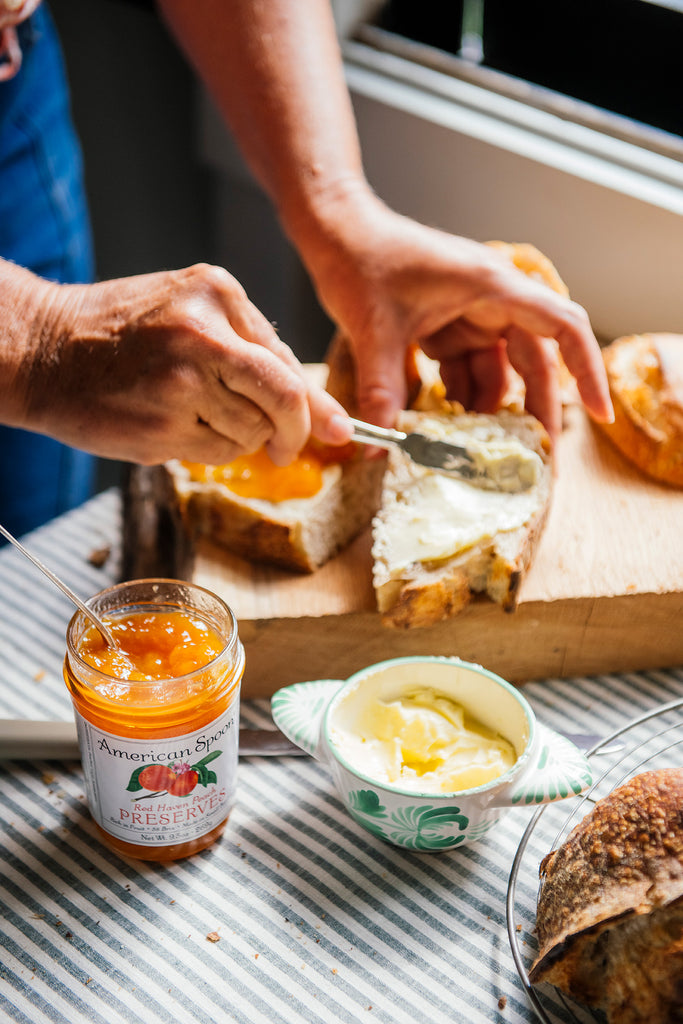 Loaves of bread with jars of Peach, Strawberry & Sour Cherry Preserves on a wooden surface