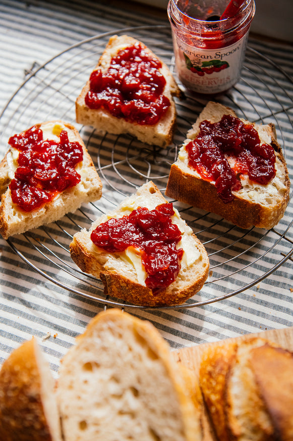 Four slices of toasted bread spread with cherry preserves on a wire cooling rack, with a jar of American Spoon Sour Cherry preserves and blurred bread slices in the foreground on a striped cloth.