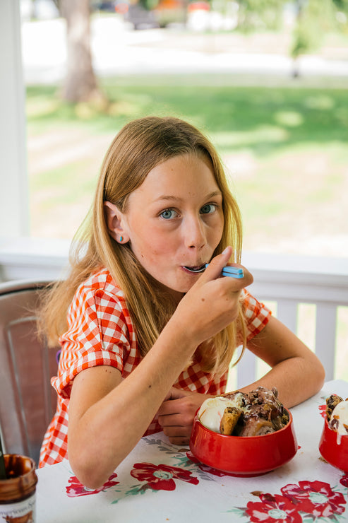 Load image into Gallery viewer, Young girl eating an ice cream sundae at a table with a floral tablecloth.
