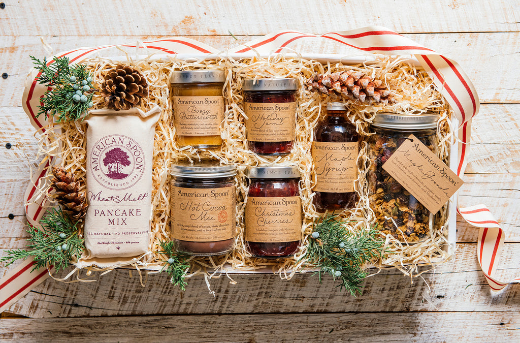 A holiday-themed gift basket filled with American Spoon preserves, granola, syrup, hot cocoa and wheat malt pancake mix, arranged in a white wooden tray with shredded paper, pinecones, evergreen branches, and a red and white striped ribbon on a white wood background.