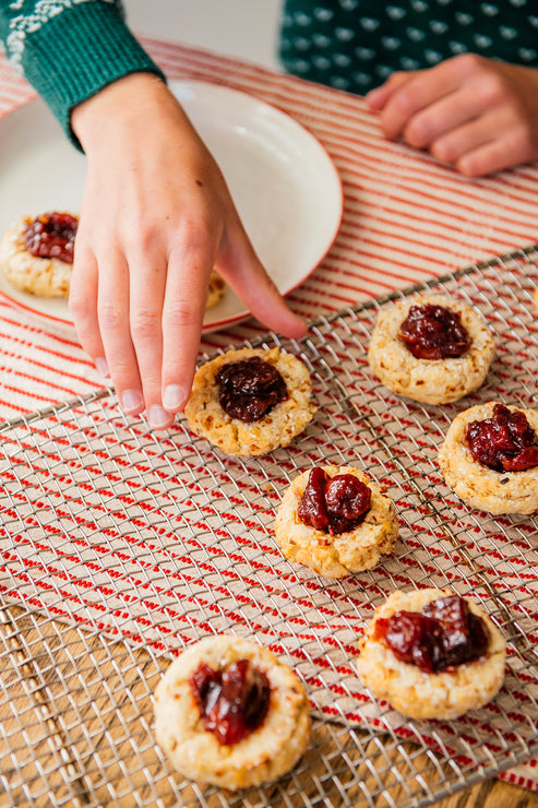 Load image into Gallery viewer, Thumbprint cookies with dark red jam centers, arranged on a wire cooling rack over a red-and-white striped cloth. A hand reaches for one cookie, with a white plate and another person's hands partially visible in the background.
