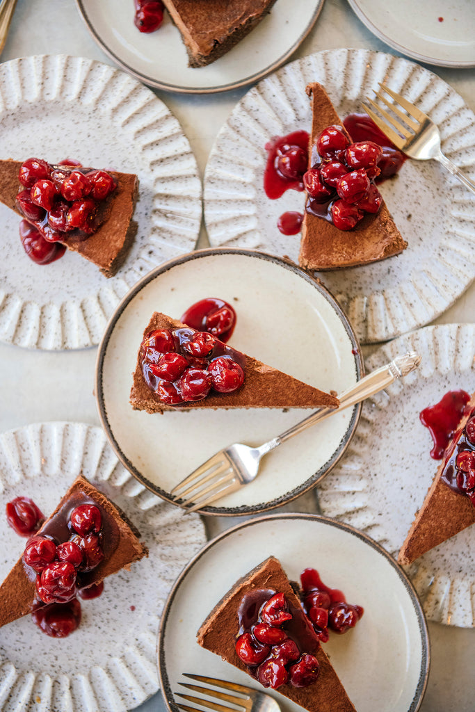 Multiple slices of chocolate cheesecake topped with fresh red cherries and berry sauce, served on speckled ceramic plates with forks, arranged on a light surface.