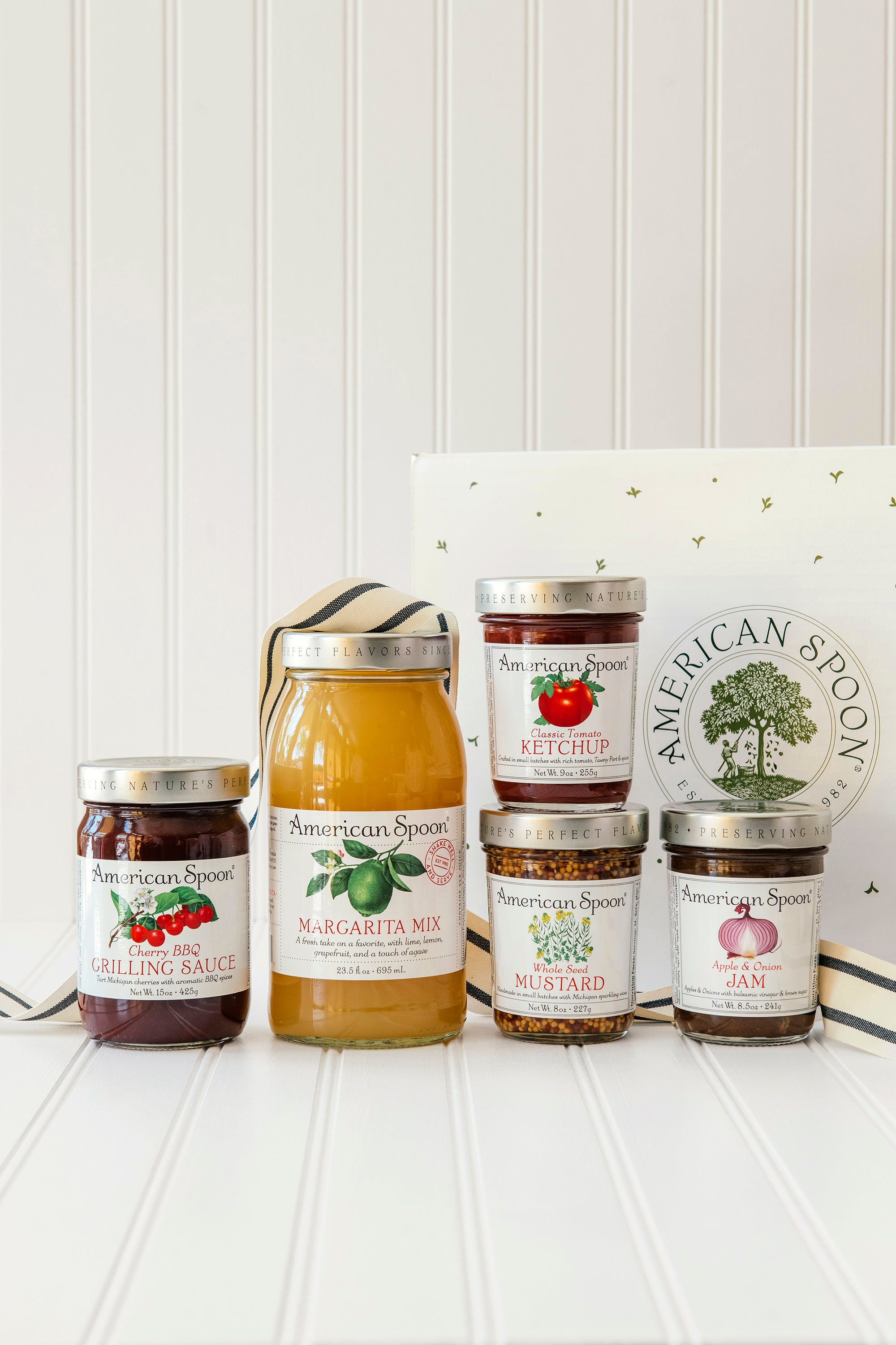 Collection of American Spoon Margarita Mix, Cherry BBQ, Classic Ketchup, Whole Seed Mustard and Apple Onion Jam on a white surface with a Gift Box