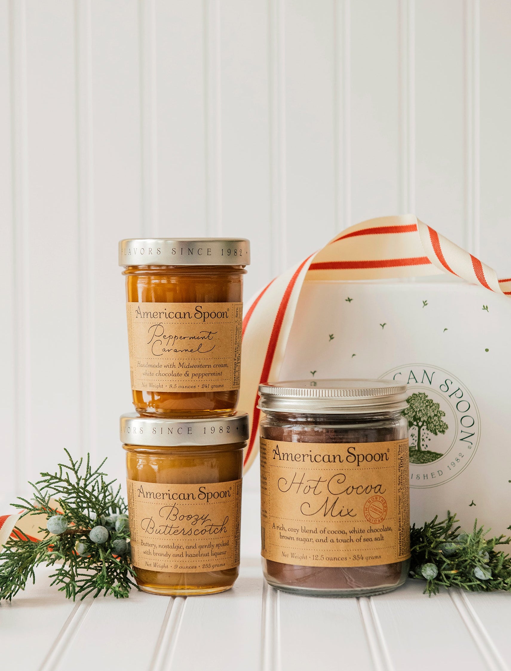 Three jars of American Spoon Hot Cocoa Mix, Boozy Butterscotch & Peppermint Caramel on a white surface with a decorative ribbon and greenery.