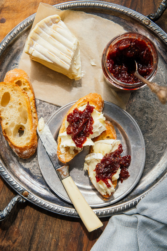 An overhead styled food scene on a large silver tray, showing a slice of white cheese, a jar American Spoon Holiday Jam with a spoon, two pieces of baguette, and two open-faced bread slices topped with cream cheese and the jam, with a knife resting on the tray.